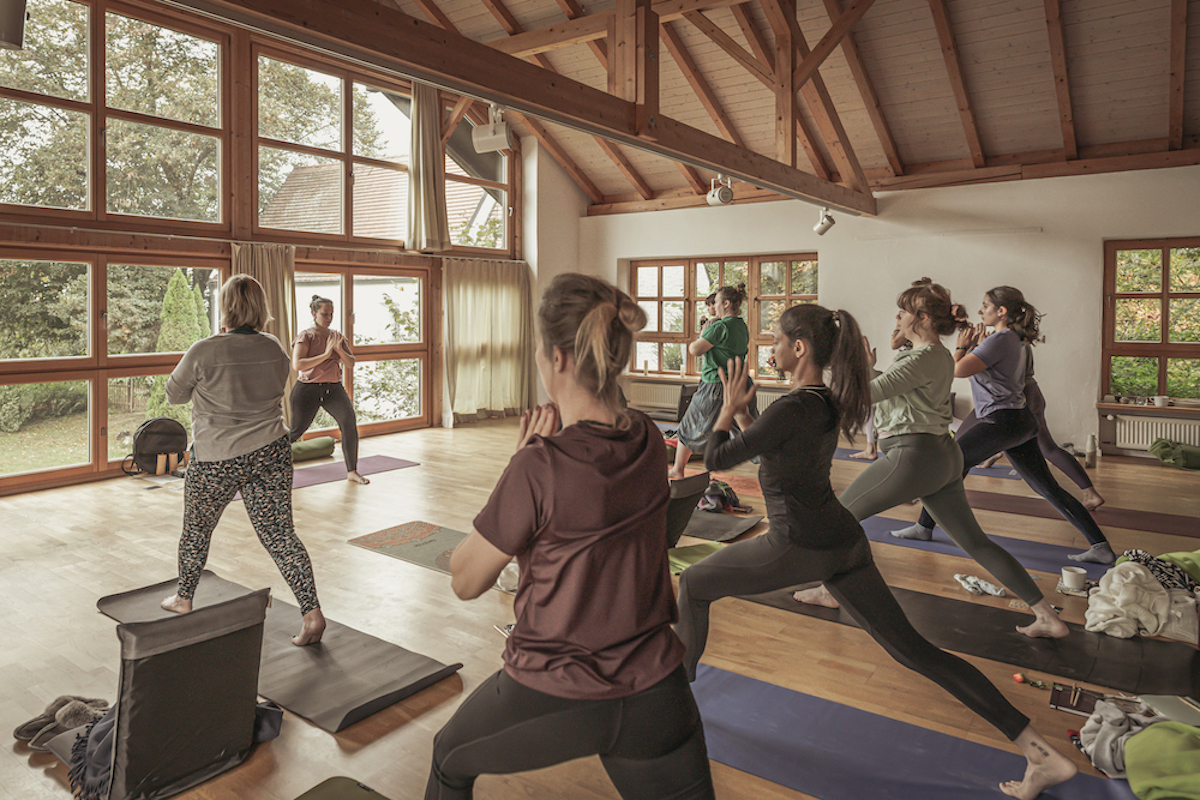 Women all practising yoga holding hands at heart in a high lunge position led by experienced guides, providing valuable tools and insights for women to prioritize self-care in their daily lives