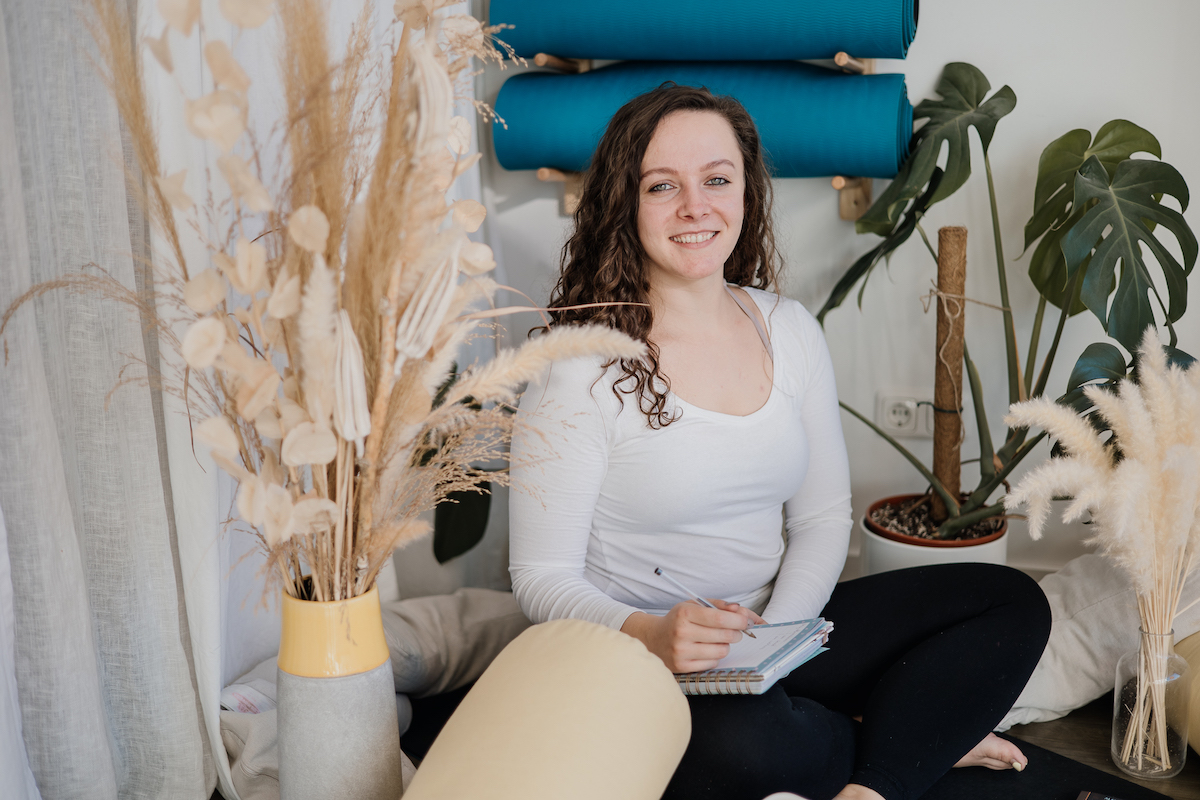 Paulina, a trauma-informed yoga teacher, smiling warmly while seated in a serene studio with plants and yoga props.