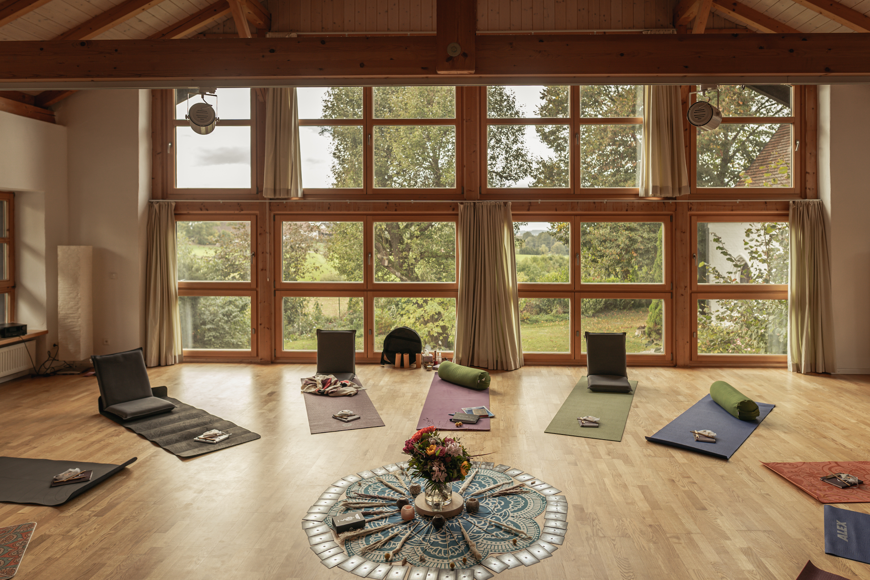Bright and inviting yoga studio with large floor-to-ceiling windows overlooking a lush green landscape. The room is set up with yoga mats, cushions, and props arranged in a circle around a beautifully decorated centerpiece featuring flowers and a mandala cloth, creating a serene and calming atmosphere.
