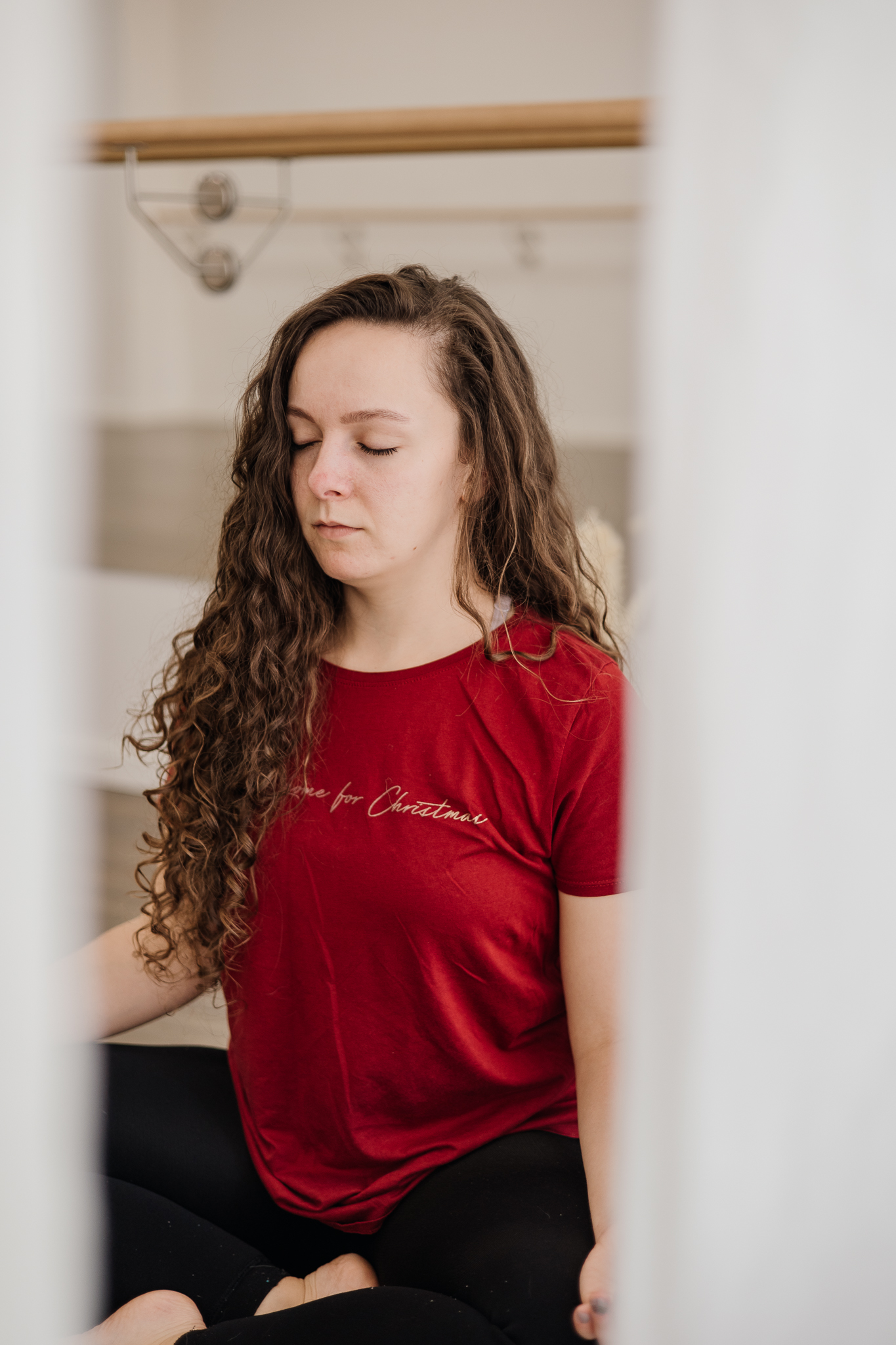 Paulina, a yoga teacher, sitting cross-legged with eyes closed in a moment of mindfulness and meditation, wearing a red shirt, in a serene studio setting.