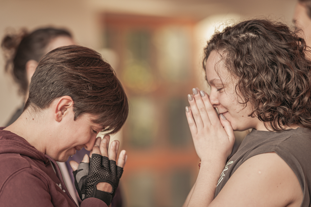 two women holding namaste, closed eyes, bowing towards each other, smiling in gratitude and compassion